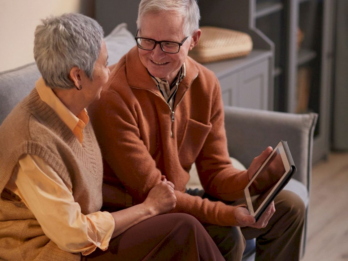 The image shows an older couple sitting on a couch, smiling at each other, with the man holding a tablet device.