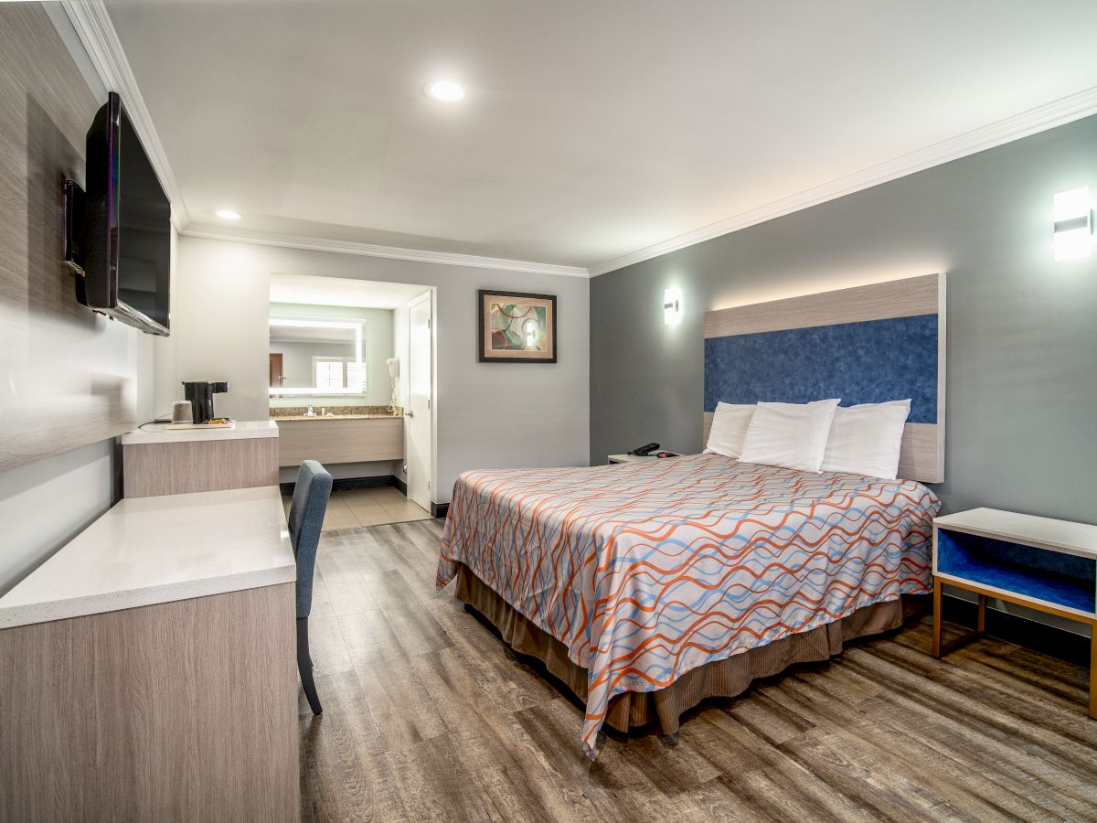 A modern hotel room with a large bed, wall-mounted TV, desk, chair, and bedside table. The room leads to a well-lit bathroom.