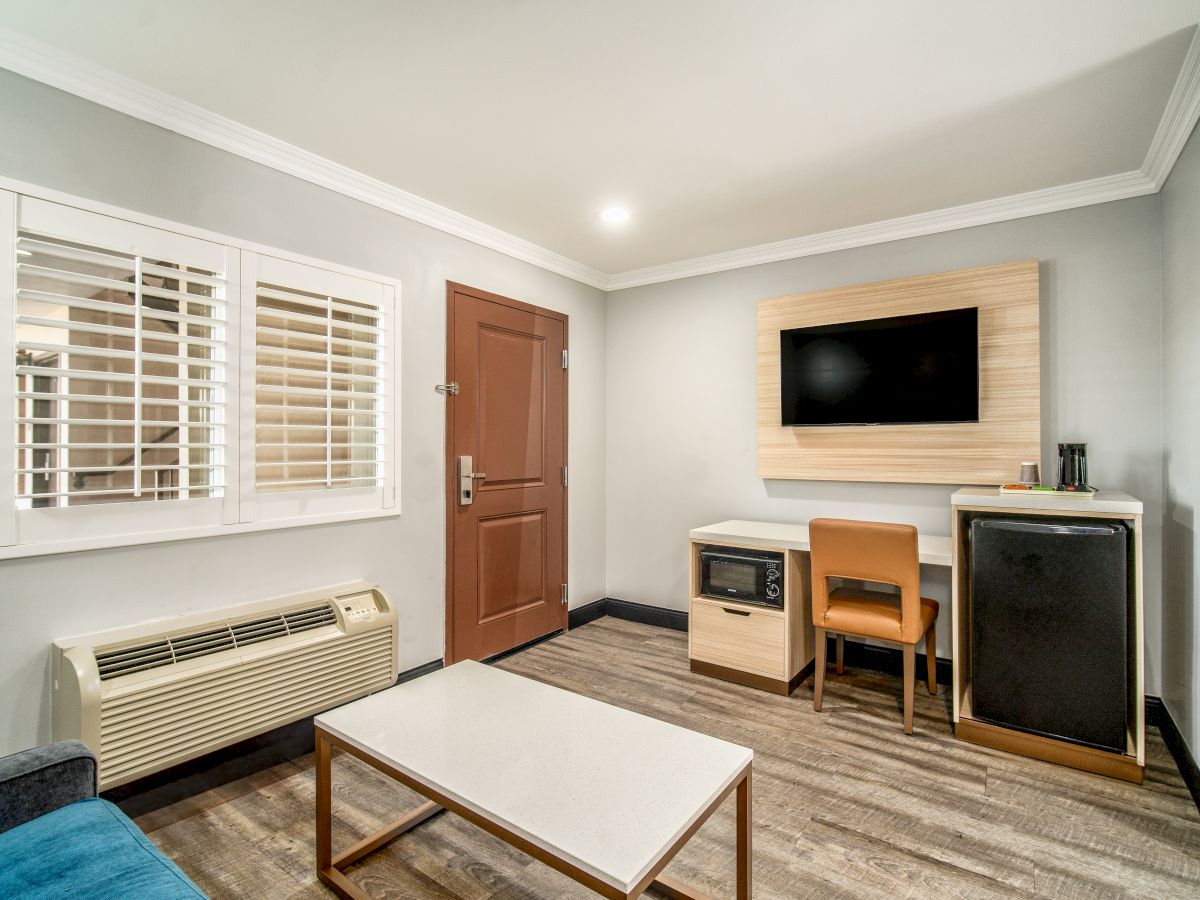 A modern room with a wall-mounted TV, table, chair, small fridge, microwave, air conditioner, coffee maker, door, window, and a coffee table.