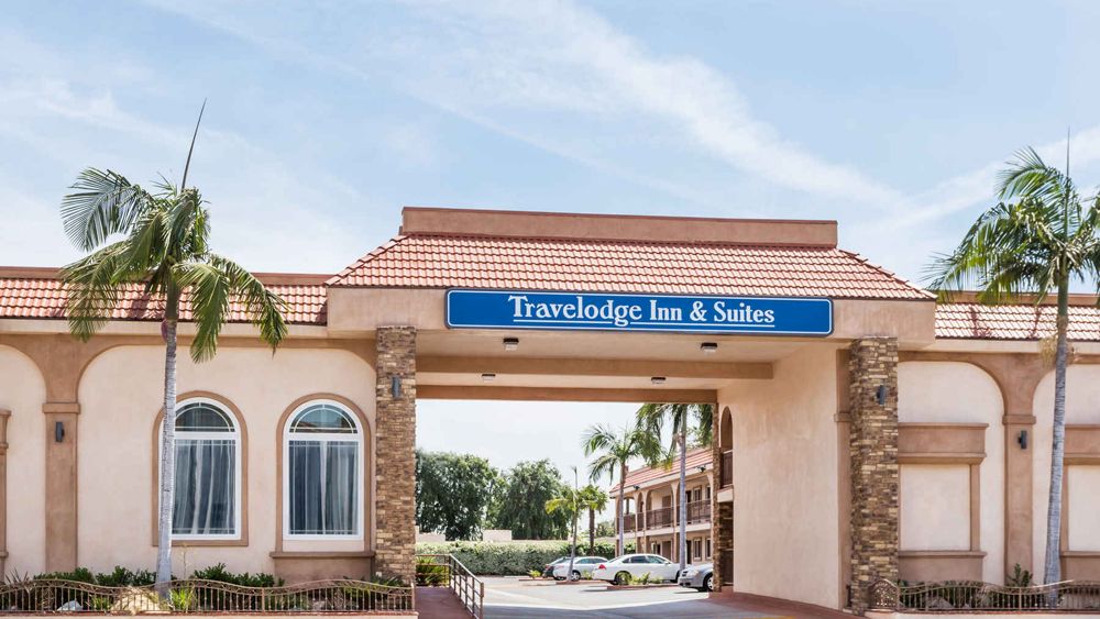 The image shows the exterior of a Travelodge Inn & Suites building with palm trees and a clear blue sky in the background.