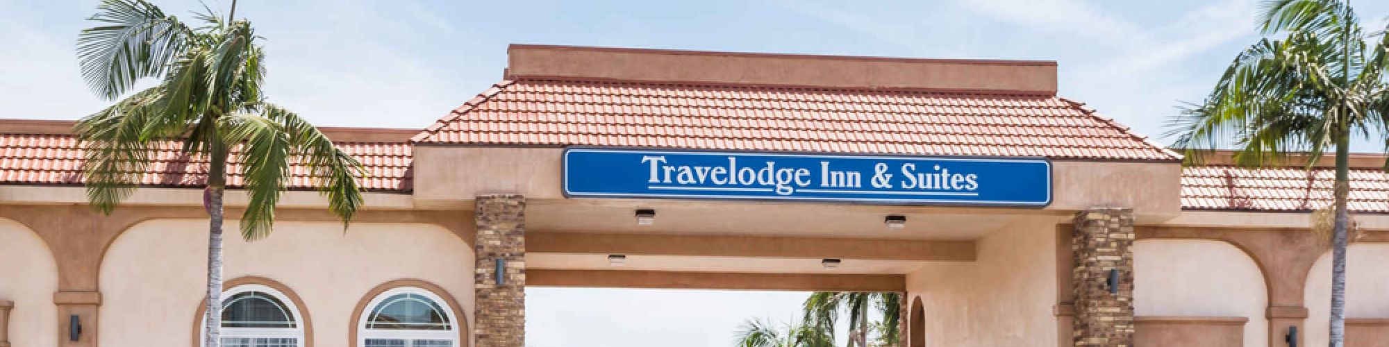 The image shows the exterior of a Travelodge Inn & Suites building with palm trees and a clear blue sky in the background.