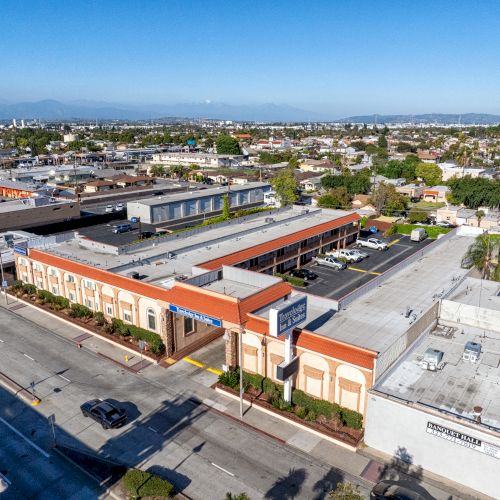 This image shows an aerial view of a suburban area with commercial buildings, a parking lot, and streets lined with vehicles, set under a clear sky.