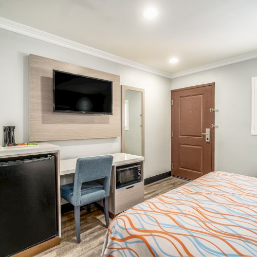 The image shows a modern hotel room with a bed, TV, desk, chair, mini-fridge, and door. The walls are light-colored, and the room is well-lit.