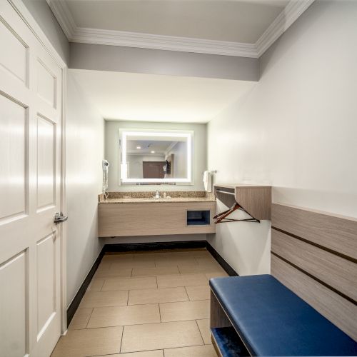 The image shows a bathroom with a white door, a large mirror with lighting, a floating vanity with a sink, and a wall bench with a blue cushion.