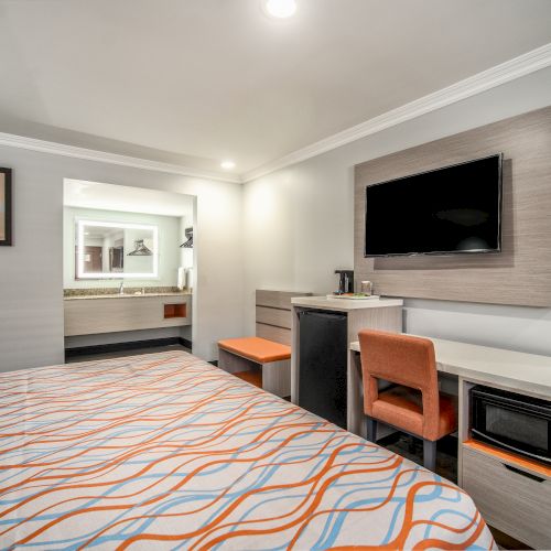 A modern hotel room with a bed, wall-mounted TV, desk, microwave, and a view of the sink and vanity area in the background.