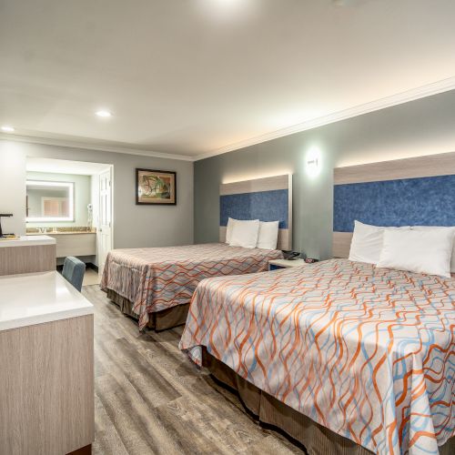 The image shows a modern hotel room with two double beds, a TV, a desk, and a bathroom in the background, featuring contemporary decor.