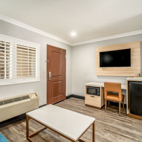 A modern hotel room featuring a door, a window with shutters, a TV, a desk and chair, a fridge, a microwave, and a wooden floor.