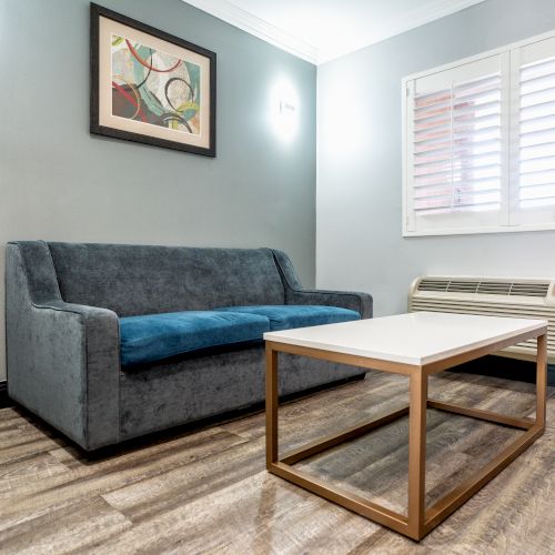 The image shows a minimalist living room with a blue sofa, a white coffee table, a wall-mounted abstract painting, and a window with blinds.