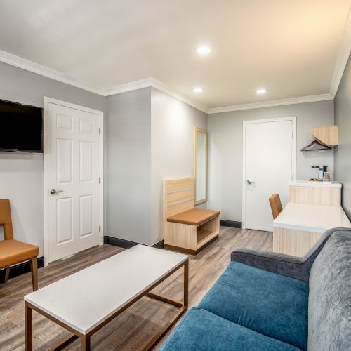 A modern living room with a blue sofa, beige chairs, a wall-mounted TV, a coffee table, and wooden flooring, featuring recessed lighting and wall decor.