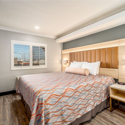 A neatly arranged hotel bedroom with a large bed, wall-mounted TV, bedside table with a phone, modern lighting, and a window with shutters.