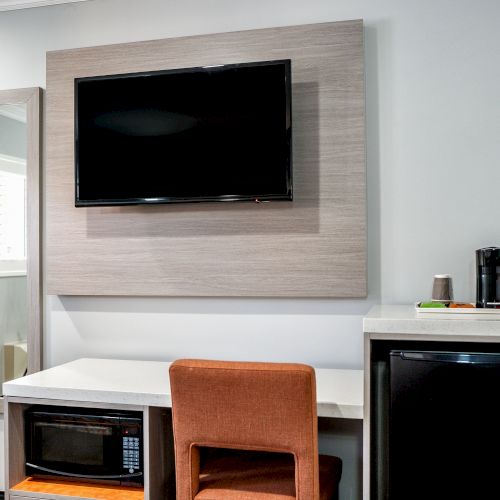 The image shows a hotel room setup with a wall-mounted TV, a mirror, a desk with a chair, a microwave, a mini-fridge, and a coffee maker.