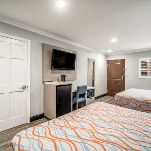 A hotel room with two beds, a wall-mounted TV, a desk with a chair, a mini-fridge, a window, and a door. Bright, minimalist design.