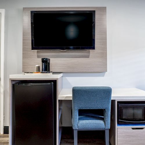 The image shows a room with a TV, mini-fridge, desk, blue chair, coffee maker, and a microwave, with a door and a window in the room.