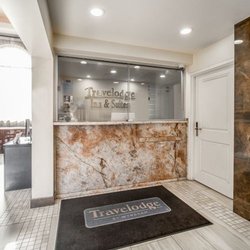 The image shows the reception area of a Travelodge Inn & Suites with a front desk, a sign, and a nearby seating area.