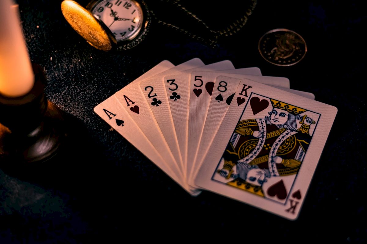 The image shows playing cards (A, 2, 3, 5, 8 of clubs and K of hearts), a lit candle, a pocket watch, and a coin on a dark surface.