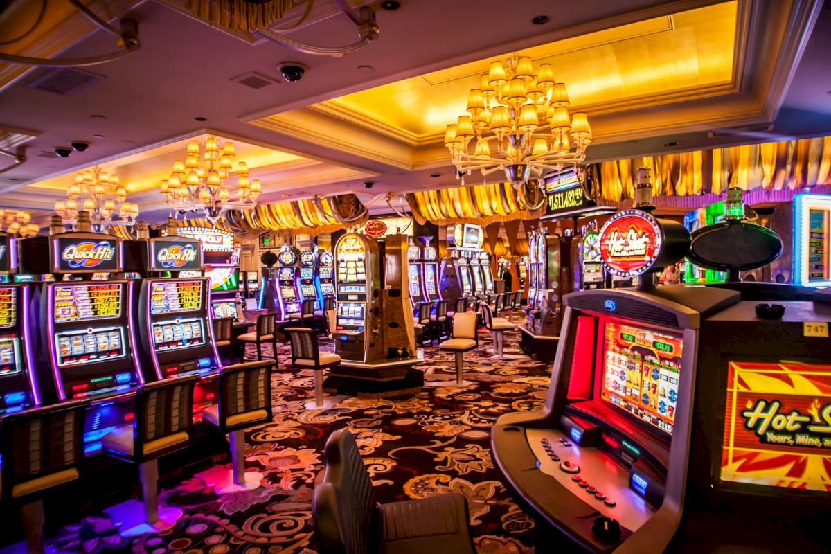 A vibrant casino gaming floor featuring numerous slot machines, bright lights, and luxurious chandeliers under a high ceiling.