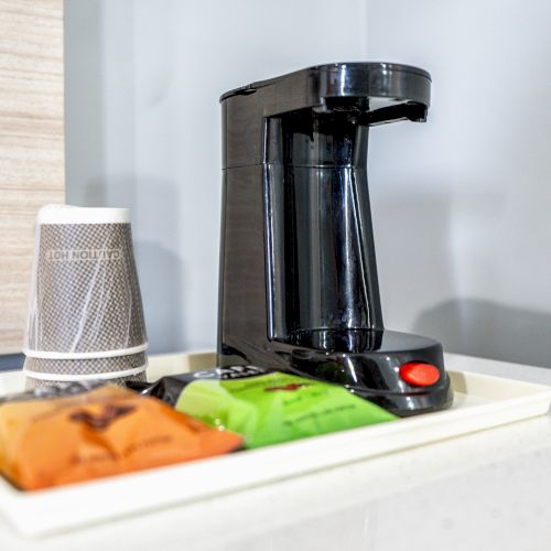 The image shows a coffee maker with a cup and three coffee packets placed on a tray against a gray and wooden background.