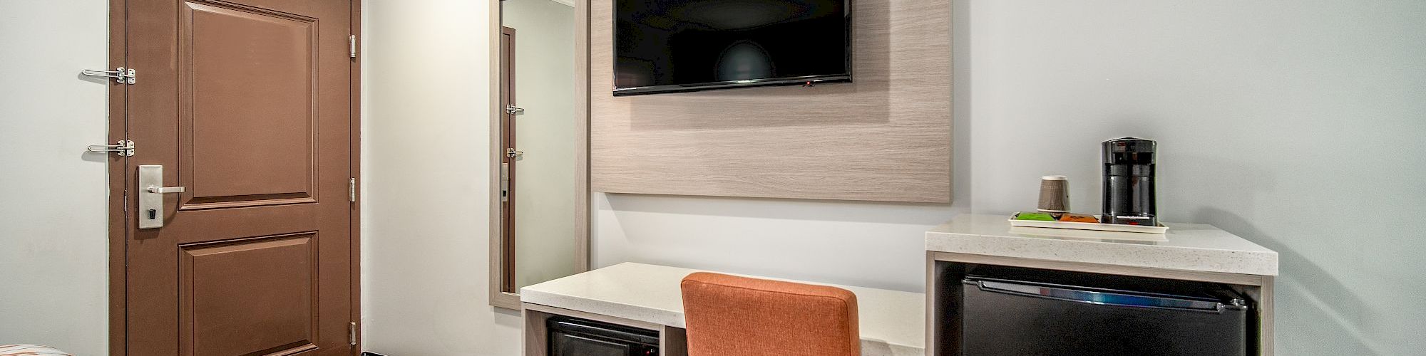 A modern hotel room corner with a brown door, wall-mounted TV, microwave, small table, coffee maker, and an orange chair.