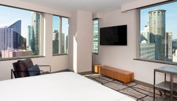 A modern hotel room with city views, a large bed, TV, armchair, and small table. The windows show skyscrapers and a clear sky outside.