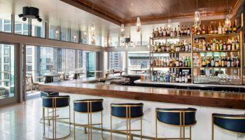 A modern bar with a long counter, high chairs, and shelves stocked with various bottles of liquor, large windows, and outdoor seating.