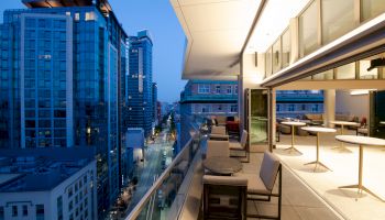 A modern cityscape at dusk with tall buildings and a sleek, open-air balcony featuring tables and chairs overlooking the city street below.