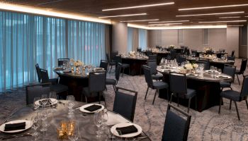 The image shows an elegantly set up banquet hall with round tables, each arranged with plates, glasses, napkins, and floral centerpieces, and chairs.