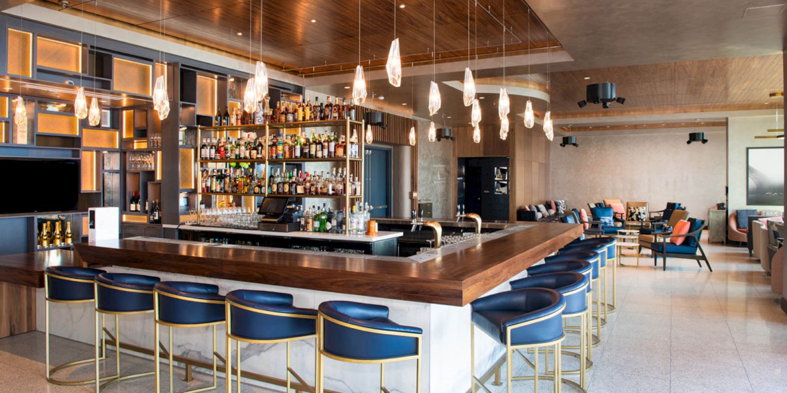 A modern bar with blue stools, a wooden countertop, hanging lights, and a well-stocked backdrop; seating area visible in the background.