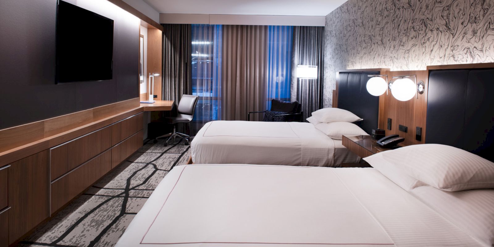 A modern hotel room with two neatly made beds, a large TV on the wall, a desk and chair near the window, and stylish decor.
