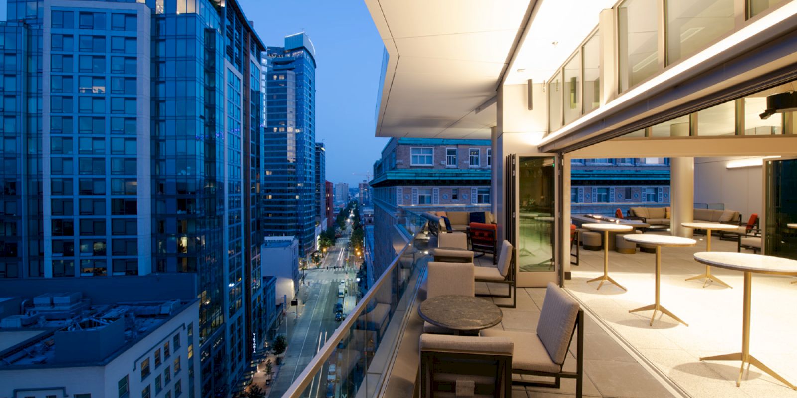 A modern balcony at dusk overlooks a city street and high-rise buildings, featuring seating areas and tables.