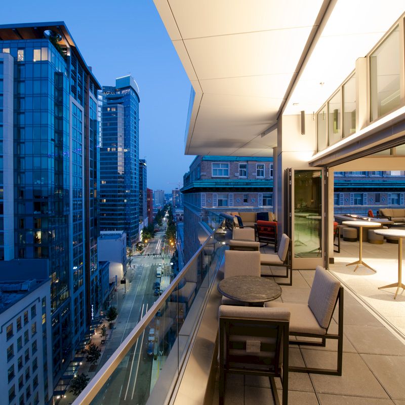 A modern urban balcony overlooking a cityscape with tall buildings, featuring outdoor seating and tables under ambient evening lighting.