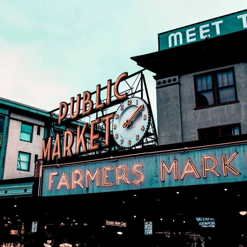 A public market sign with a clock, and a bright neon "Farmers Market" sign below it, attached to a building with other visible structures.