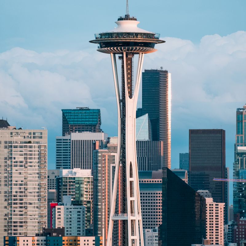 The image shows the Space Needle towering over the city skyline, with various skyscrapers and buildings in the background, under a partly cloudy sky.