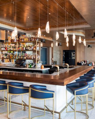 A modern bar with blue padded stools, a wooden counter, and pendant lights. It has a well-stocked display of bottles and a seating area in the background.