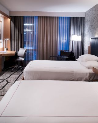 A modern hotel room with two neatly made beds, a wall-mounted TV, a work desk with a chair, and contemporary decor.