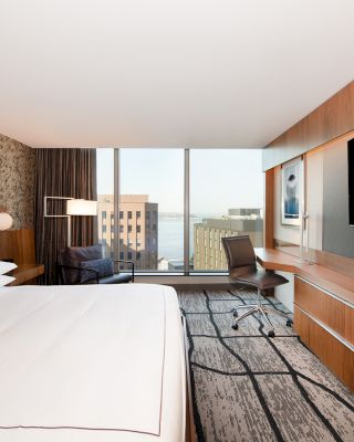 A modern hotel room with a large bed, a desk, a chair, a wall-mounted TV, and a view of the city through a large window.