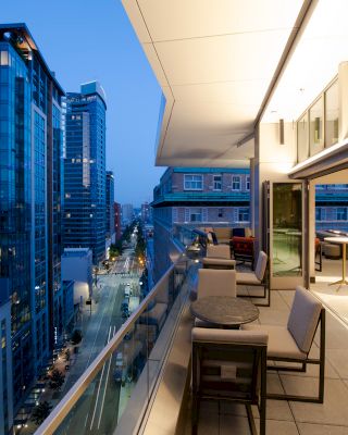 A modern urban balcony at dusk with seating, tables, and a glass railing overlooking a city street with tall buildings.