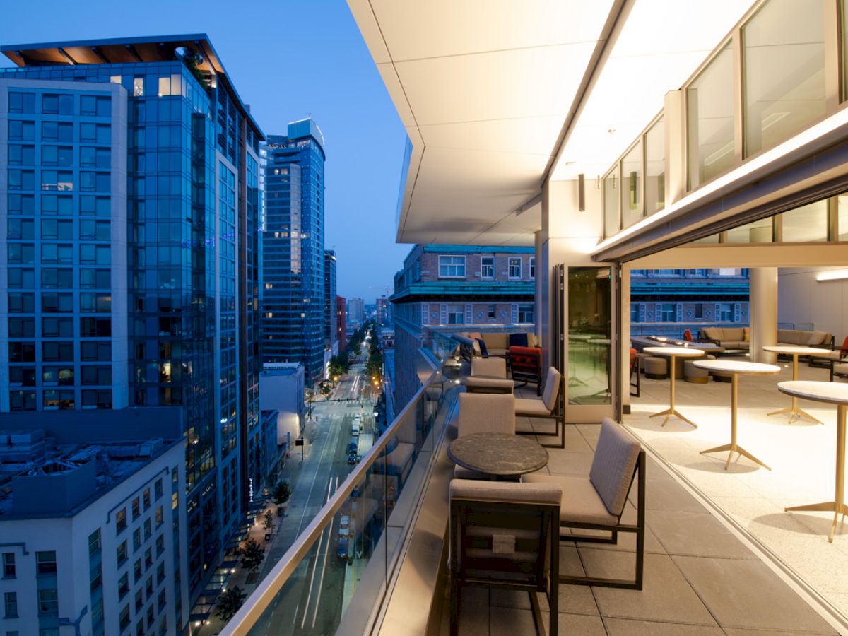 This image features a modern rooftop terrace with seating and a cityscape view, showcasing tall buildings and a lighted street.