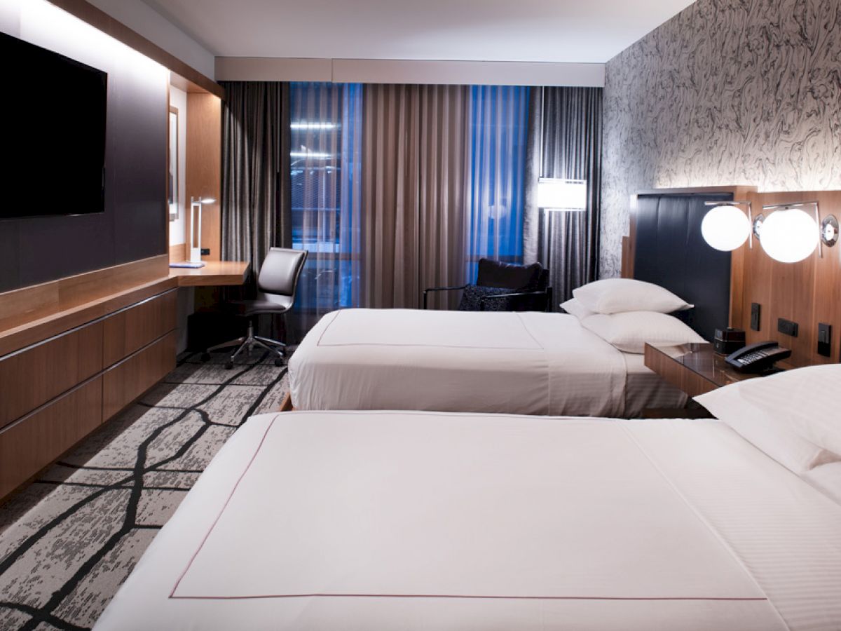 The image shows a modern hotel room with two neatly made beds, a large TV, a desk with a chair, and contemporary lighting and design elements.