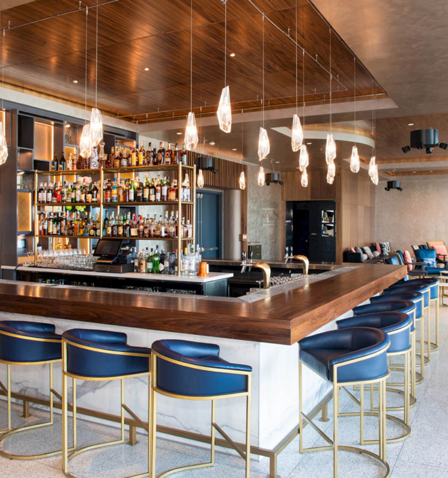 The image shows a modern bar with blue stools, a well-stocked liquor shelf, hanging pendant lights, and comfortable seating in the background.