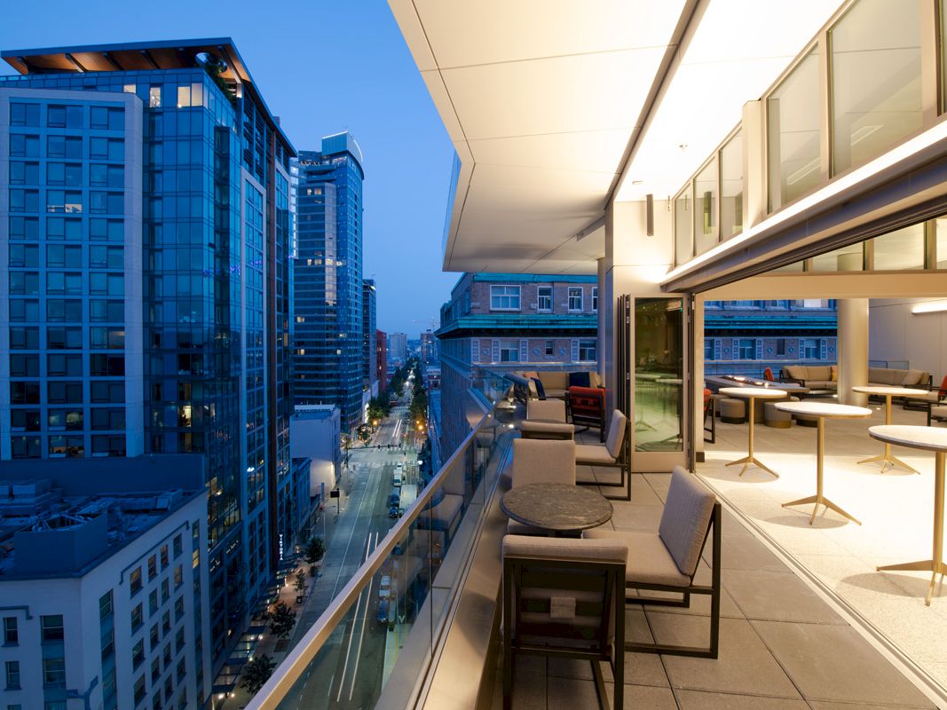 The image shows a modern balcony with seating and tables, overlooking a city with tall buildings during evening time.