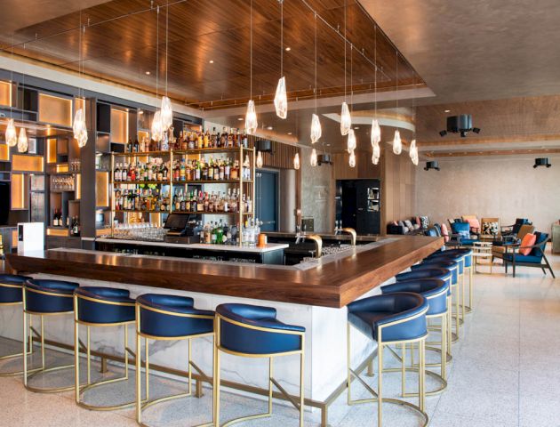 A modern bar with blue chairs, a well-stocked liquor shelf, pendant lighting, and a spacious seating area with tables and chairs in the background.