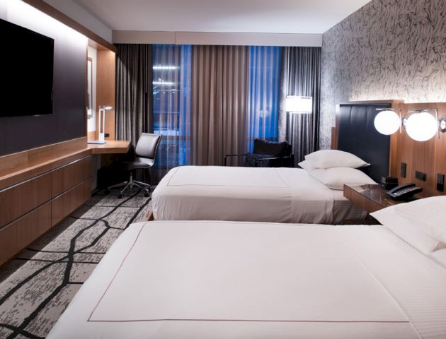The image shows a modern hotel room with two neatly made beds, a TV, a desk with a chair, and stylish lighting.