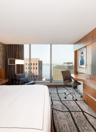 A modern hotel room with a large bed, wall-mounted TV, desk, and chair, overlooking a cityscape with buildings and water through wide windows.