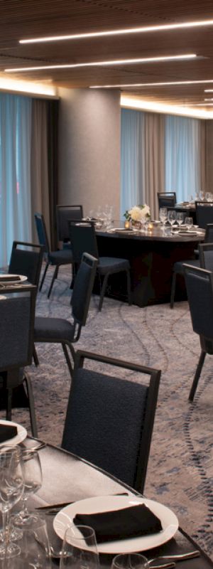 The image depicts an elegant banquet hall set up with round tables covered in dark tablecloths, each arranged with tableware, glasses, and floral centerpieces.