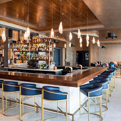 This image shows a modern bar with a central counter, blue cushioned stools, hanging pendant lights, and shelves stocked with bottles in the background.