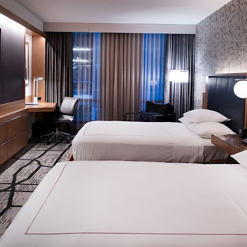 A modern hotel room features two beds, a flat-screen TV, a desk with a chair, stylish decor, wood furnishings, and large windows with curtains.