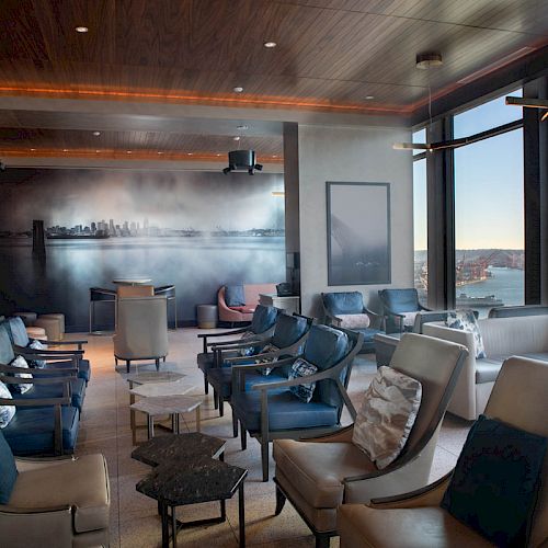 A modern, cozy lounge with ample seating, large windows offering a scenic view of water and a Ferris wheel, and a cityscape mural on the wall.