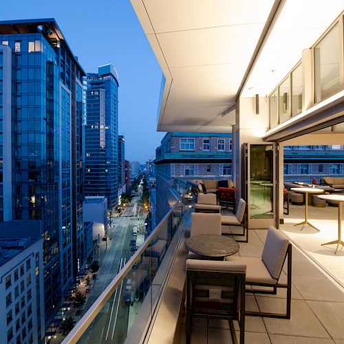 A modern outdoor balcony with seating, overlooking a cityscape of tall buildings at twilight, featuring large glass windows and tables.