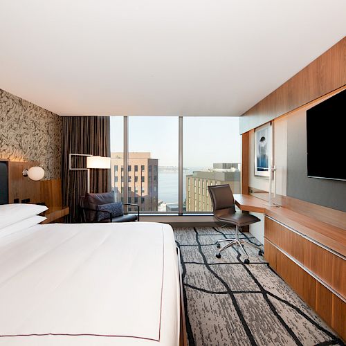 A modern hotel room featuring a large bed, wall-mounted TV, desk with chair, and a window with a cityscape view.