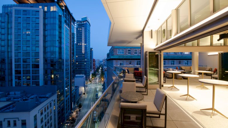 The image shows a modern cityscape viewed from a balcony with seating and tables at dusk. Tall buildings and a street visible below.